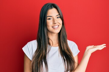 Young brunette woman wearing casual white tshirt over red background smiling cheerful presenting and pointing with palm of hand looking at the camera.