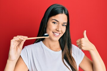 Young brunette woman holding toothbrush with toothpaste smiling happy and positive, thumb up doing excellent and approval sign