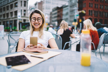 Portrait of happy Asian student in classic eyewear smiling at camera while using cellphone for research education information, cheerful hipster girl with smartphone technology posing at street cafe