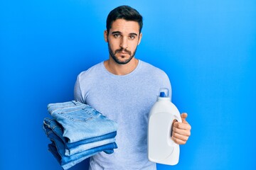 Young hispanic man holding folded jeans and detergent bottle skeptic and nervous, frowning upset...