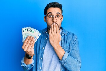 Young hispanic man holding bunch of 20 euro banknotes covering mouth with hand, shocked and afraid for mistake. surprised expression