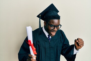 Handsome black man wearing graduation cap and ceremony robe holding diploma screaming proud,...