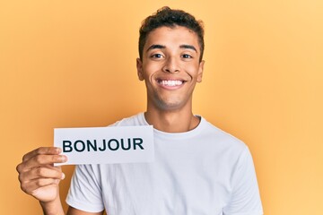 Young handsome african american man holding bonjour french greeting word looking positive and happy standing and smiling with a confident smile showing teeth © Krakenimages.com