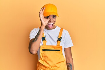 Young handsome african american man wearing handyman uniform over yellow background doing ok gesture with hand smiling, eye looking through fingers with happy face.