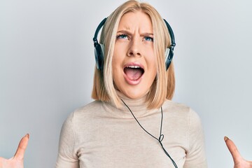 Young blonde woman listening to music using headphones crazy and mad shouting and yelling with aggressive expression and arms raised. frustration concept.