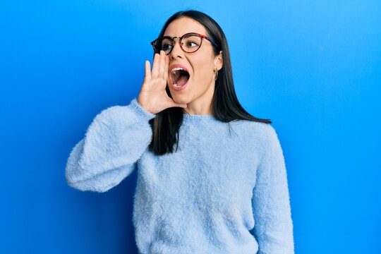 Young brunette woman wearing casual clothes and glasses shouting and screaming loud to side with hand on mouth. communication concept.