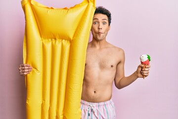Obraz premium Young handsome man wearing swimsuit holding summer mattress float and icecream making fish face with mouth and squinting eyes, crazy and comical.