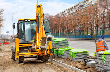 An orange road excavator works at a work site paving paving slabs along a city street on a spring afternoon.