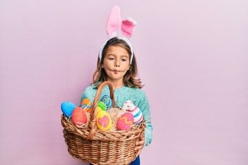 Little beautiful girl wearing cute easter bunny ears holding wicker basket with colored eggs making fish face with mouth and squinting eyes, crazy and comical.