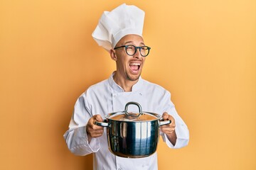 Bald man with beard wearing professional cook holding cooking pot angry and mad screaming frustrated and furious, shouting with anger. rage and aggressive concept.