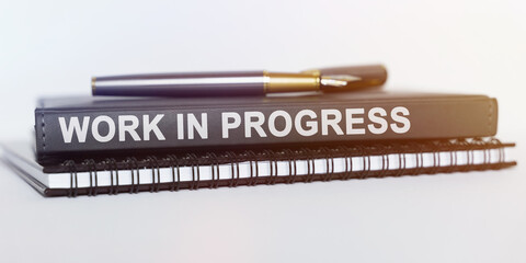 On the table are a notebook, a pen and a book. The book says - WORK IN PROGRESS