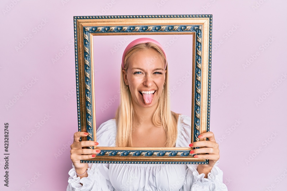 Poster Young blonde girl holding empty frame sticking tongue out happy with funny expression.