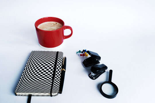 A business persons working space. A checkered notebook , a magnifying glass , Ink pen, staple, some pins and a red coffee mug beside them.