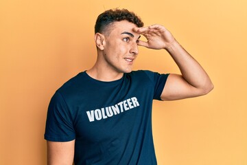 Hispanic young man wearing volunteer t shirt very happy and smiling looking far away with hand over head. searching concept.