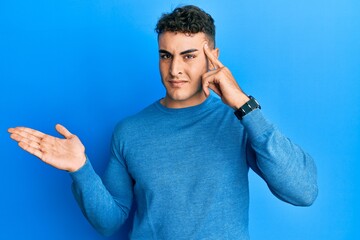 Hispanic young man wearing casual winter sweater confused and annoyed with open palm showing copy space and pointing finger to forehead. think about it.