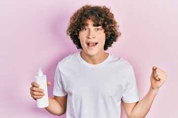 Handsome young man holding cosmetic moisturizer facial cream screaming proud, celebrating victory and success very excited with raised arms