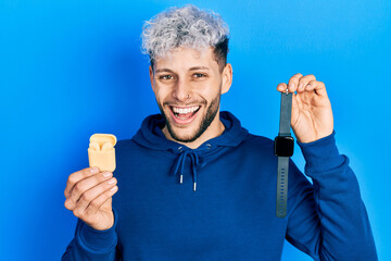 Young hispanic man with modern dyed hair holding wireless earphones and smart watch smiling and...
