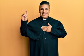 Young latin priest man standing over yellow background smiling swearing with hand on chest and...