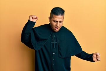 Young latin priest man standing over yellow background dancing happy and cheerful, smiling moving...