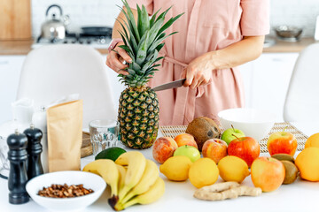 Woman cutting pineapple. Composition of different fruits and vegetables on white light kitchen.