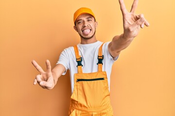 Hispanic young man wearing handyman uniform smiling with tongue out showing fingers of both hands doing victory sign. number two.