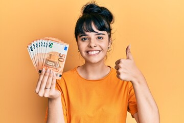 Young hispanic girl holding bunch of 50 euro banknotes smiling happy and positive, thumb up doing...