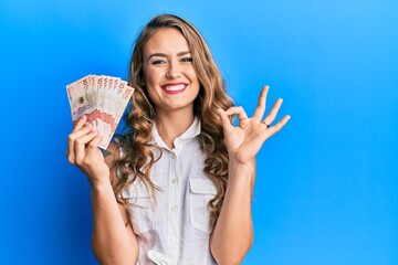 Young blonde girl holding 10 colombian pesos banknotes doing ok sign with fingers, smiling friendly gesturing excellent symbol