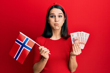 Young hispanic woman holding norway flag and krone banknotes puffing cheeks with funny face. mouth inflated with air, catching air.