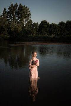 Conceptual fine art portrait of a young girl standing in the water of lake in pink dress holding bouquet of flowers like a nymph