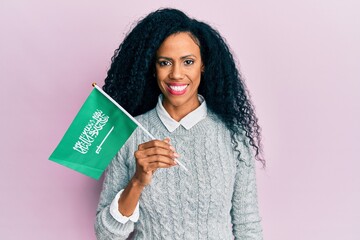 Middle age african american woman holding kingdom of saudi arabia flag looking positive and happy standing and smiling with a confident smile showing teeth