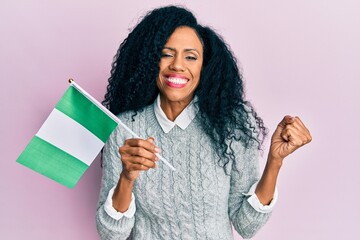 Middle age african american woman holding nigeria flag screaming proud, celebrating victory and success very excited with raised arm