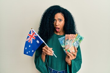 Middle age african american woman holding australian flag and dollars in shock face, looking skeptical and sarcastic, surprised with open mouth