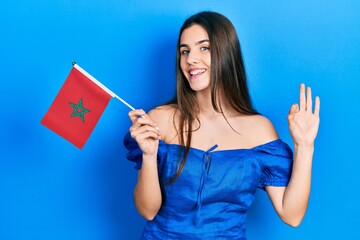 Young brunette teenager holding morocco flag doing ok sign with fingers, smiling friendly gesturing excellent symbol