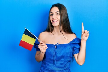 Young brunette teenager holding belgium flag smiling with an idea or question pointing finger with happy face, number one