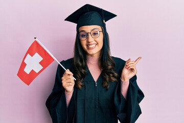 Young hispanic woman wearing graduation uniform holding switzerland flag smiling happy pointing with hand and finger to the side