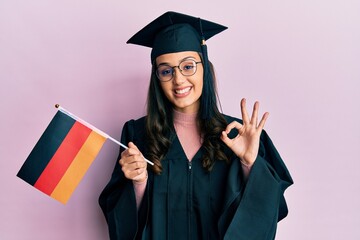 Young hispanic woman wearing graduation uniform holding germany flag doing ok sign with fingers, smiling friendly gesturing excellent symbol
