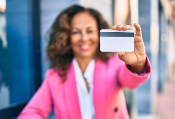 Middle age hispanic businesswoman smiling happy holding credit card standing at the city.
