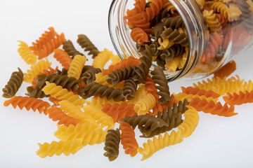 Glass jar with colorful spiral pasta falling out of it on white background
