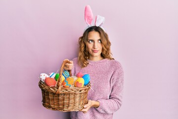 Young caucasian woman wearing cute easter bunny ears holding colored egg smiling looking to the...