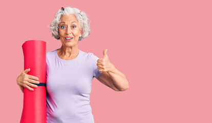 Senior grey-haired woman holding yoga mat smiling happy and positive, thumb up doing excellent and...