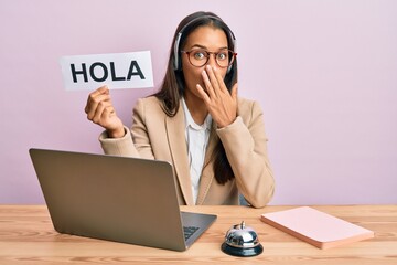 Beautiful hispanic woman wearing operator headset holding hola greenting covering mouth with hand, shocked and afraid for mistake. surprised expression