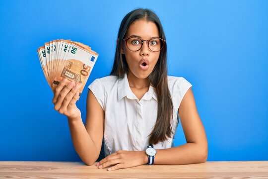 Beautiful hispanic woman holding bunch of 50 euro banknotes scared and amazed with open mouth for surprise, disbelief face
