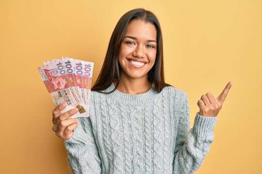 Beautiful hispanic woman holding 100 hong kong dollars banknotes smiling happy pointing with hand and finger to the side