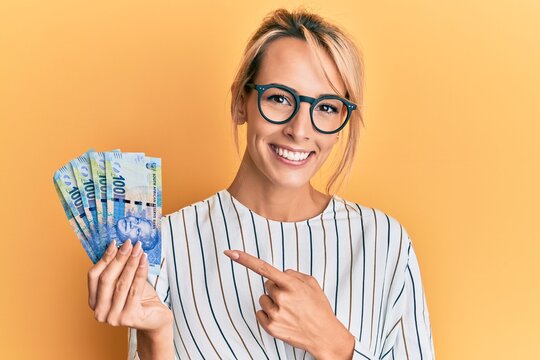 Beautiful blonde woman holding south african 100 rand banknotes smiling happy pointing with hand and finger