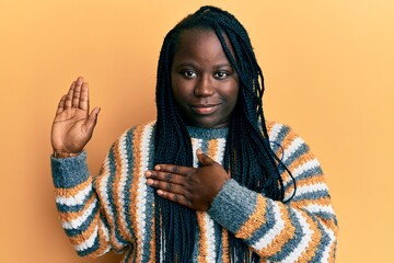 Young black woman with braids wearing casual winter sweater swearing with hand on chest and open...