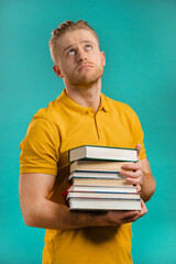 European student is dissatisfied with amount of homework and books on blue background. Man sighs and rolls his eyes in displeasure, he is annoyed, discouraged frustrated by studies.