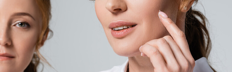 Cropped view of woman applying face cream near friend on blurred background isolated on grey, banner