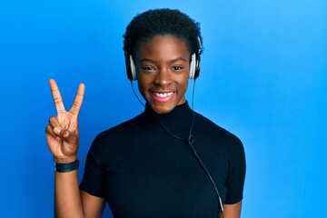 Young african american girl listening to music using headphones smiling with happy face winking at...