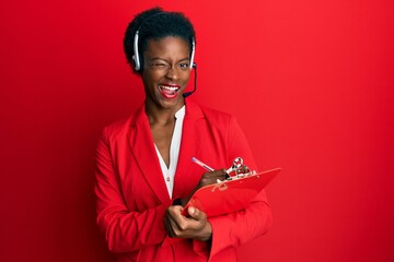 Young african american girl wearing call center agent headset writing on clipboard winking looking...