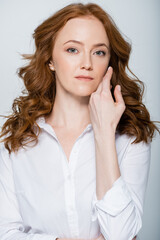 Red haired woman with hand near face looking at camera isolated on grey